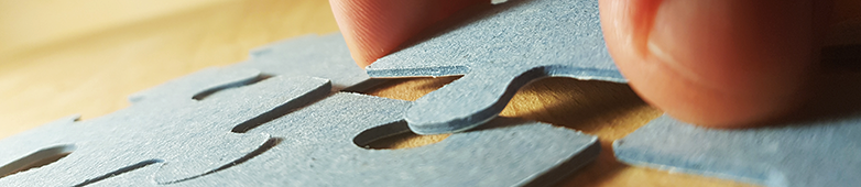 Das Bild zeigt die Detailaufnahme eines Puzzles auf einem Tisch, in das eine Hand gerade ein neues Bauteil einsetzt.